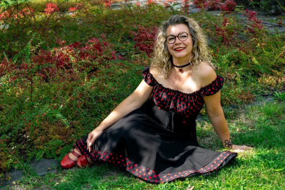 Femme assise dans une robe noire à pois rouges, entourée de plantes colorées.