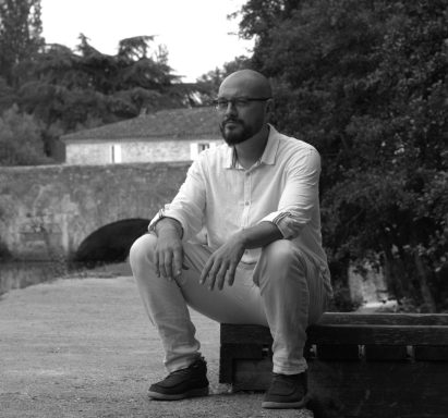 Homme assis au bord de l'eau, portant une chemise claire et des lunettes, en noir et blanc.