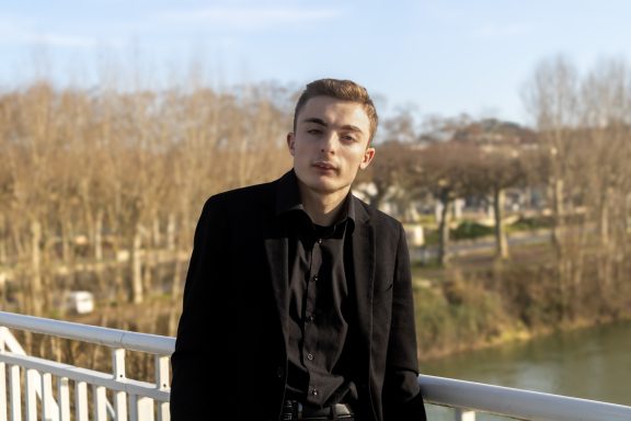 Jeune homme en noir, posé sur un pont avec un paysage naturel en arrière-plan.